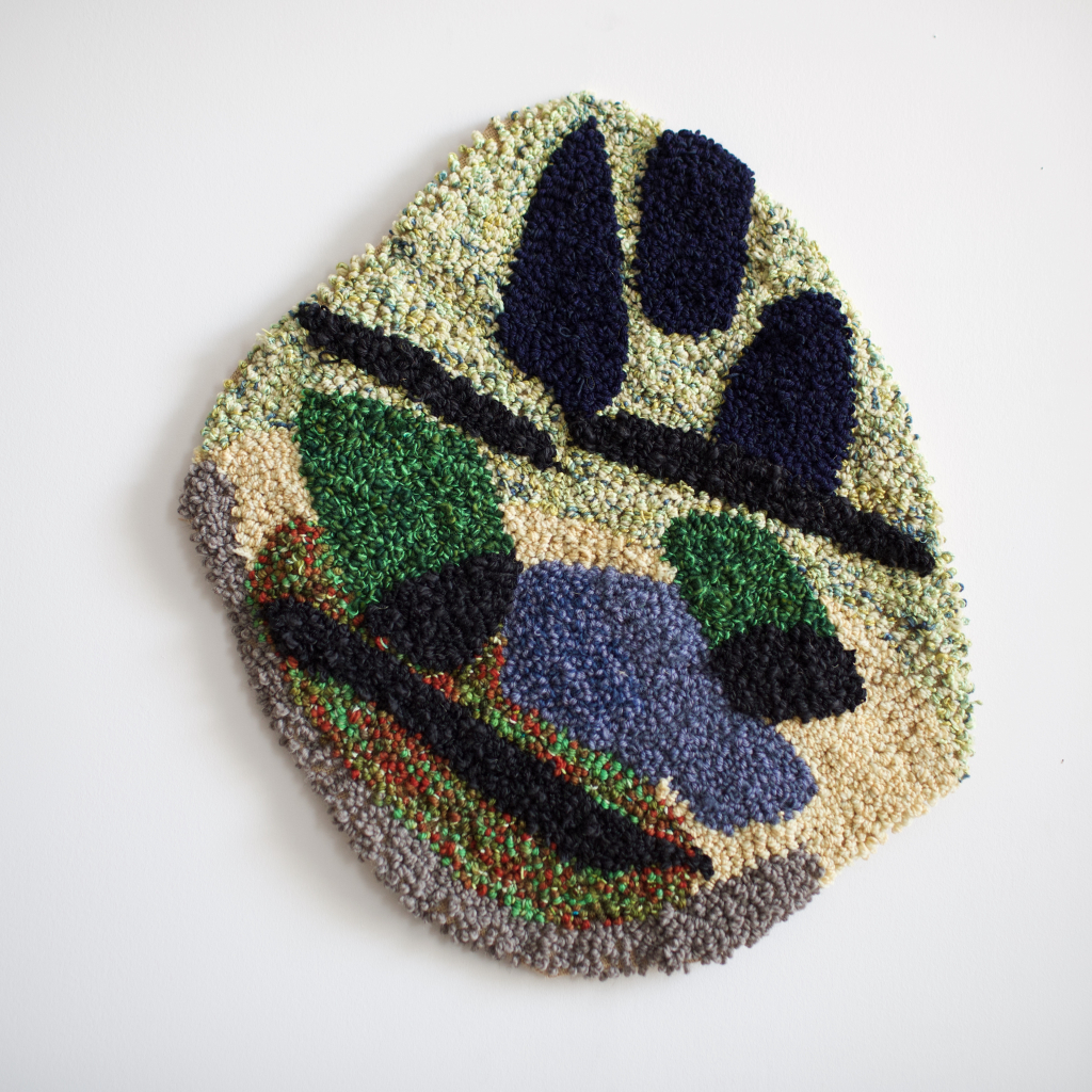 Punch needle wall piece that is round and is shades of green. It is an abstract face.
