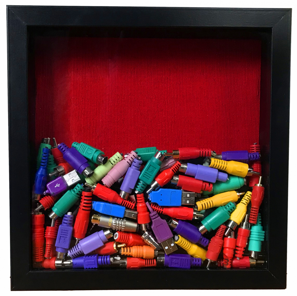 Black shadow box half filled with bright coloured plugs from computer, audio and video cables. Red fabric background.