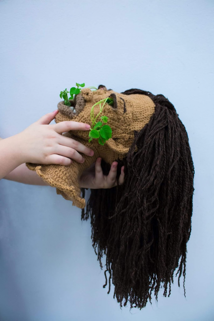 A crocheted head with green leaves sprouting from the mouth and eye. The head is tilted back with a neutral expression.